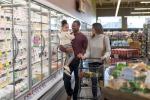 Dad holds his pre-school age daughter at the grocery. Mom is next to him pushing the cart. They are both chuckling and looking at their daughter affectionately as they stroll through the refrigerated dairy section.